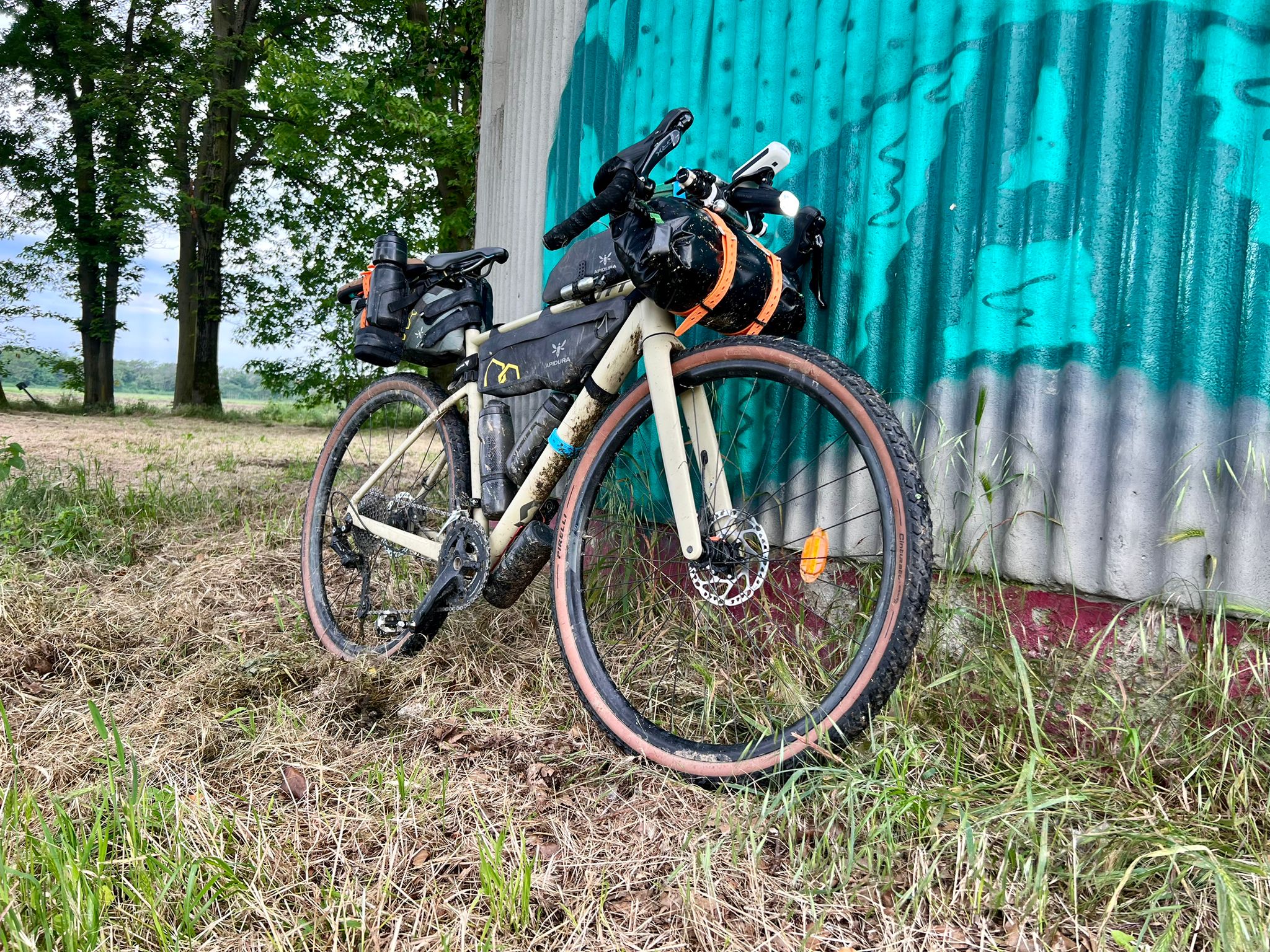 Scott Speedster Gravel bicycle, beige, with bikepacking bags attached to handlebar, frame and saddle. Bike is leaning on a abandoned industrial building wall with a graffiti in a field.